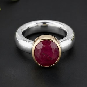 Op de afbeelding: Een ring met een dikke, gepolijste zilveren band en een ronde, gefacetteerde dieprode edelsteen in een goudkleurige zetting. De steen is in het midden van de band geplaatst, wat zorgt voor een contrast tussen het zilver, het goud en de rode kleur.