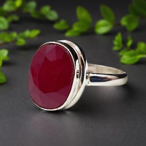 Pode incluir: Um anel de prata com uma grande pedra preciosa vermelho-escura, de formato oval e facetada, engastada numa moldura de metal polido. O aro é liso e metálico, apresentando um design limpo que destaca o contraste entre a pedra e a prata.