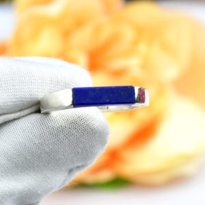 Op de afbeelding: Een zilveren ring met een rechthoekige, diepblauwe lapis lazuli steen. De ring wordt vastgehouden door een gehandschoende hand tegen een vage oranje en groene achtergrond. De steen is ongeveer 1,3 cm lang.