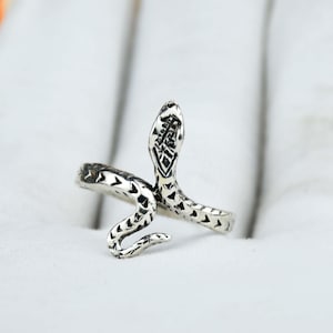May include: A silver-toned snake ring with a detailed design. The snake's head is raised, and the body wraps around the finger. The ring has a textured pattern and is set against a soft, white background. A unique piece of jewelry.