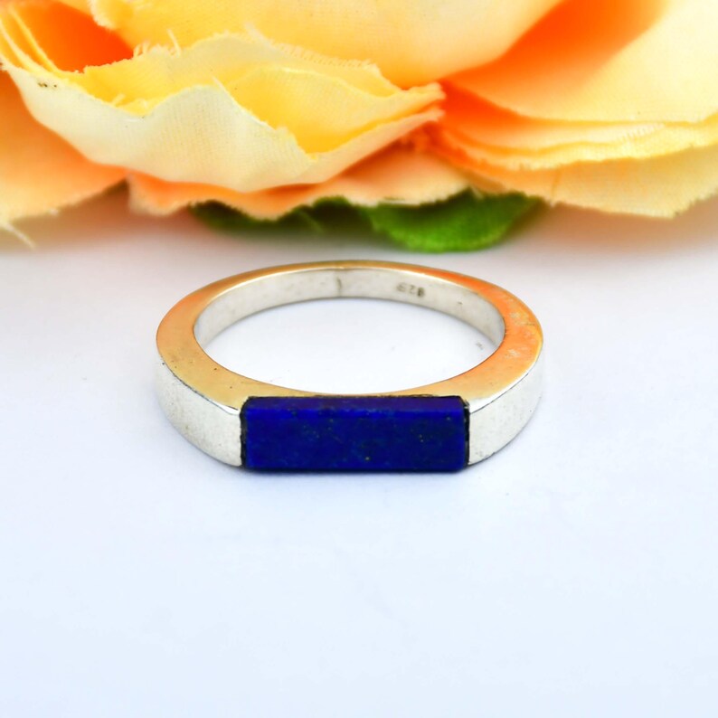 Op de afbeelding: Een zilveren ring met een rechthoekige, diepblauwe steen in het midden. De ring is geplaatst tegen een zachte, vage achtergrond van gele en oranje bloemblaadjes. Een eenvoudig, elegant sieraad.
