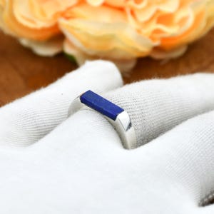 Op de afbeelding: Een zilveren ring met een rechthoekige, diepblauwe steen. De ring ligt op een witte ondergrond, met een vage achtergrond van gele en oranje bloemen. Het ontwerp van de ring is eenvoudig en modern, geschikt voor dagelijks gebruik.