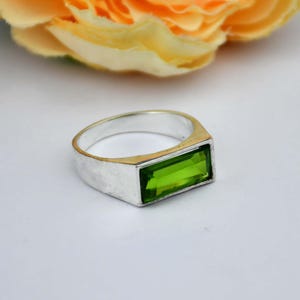 May include: A silver ring with a rectangular, bright green gemstone. The ring has a gold-colored band and a silver-colored face. The ring is set against a white background with a blurred yellow flower in the background.