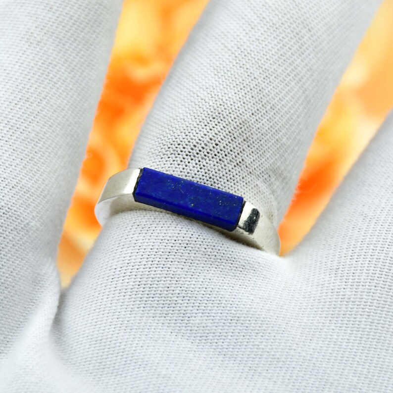 Op de afbeelding: Een zilveren ring met een rechthoekige, diepblauwe steen. De ring heeft een eenvoudig, modern ontwerp, waarbij de steen gelijk met de band is geplaatst. De ring wordt getoond op een witte ondergrond.