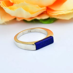 Op de afbeelding: Zilveren ring met een rechthoekige, diepblauwe lapis lazuli stenen inleg. De ring heeft een modern, minimalistisch design met een gepolijste afwerking. De ring staat tegen een zachte, vage achtergrond van gele en oranje bloemen.