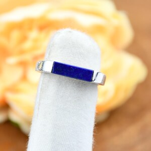 Op de afbeelding: Een zilveren ring met een rechthoekige, diepblauwe lapis lazuli steen in het midden. De ring wordt gepresenteerd op een wit oppervlak, met een vage gele en bruine achtergrond.