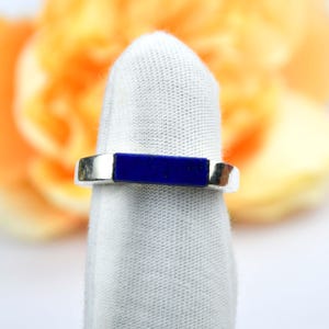 Op de afbeelding: Een zilveren ring met een rechthoekige, diepblauwe steen in het midden. De ring wordt gepresenteerd op een witte stof, met een vage oranje en gele achtergrond.