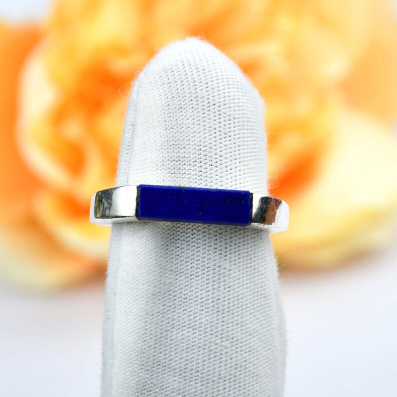 Op de afbeelding: Een zilveren ring met een rechthoekige, diepblauwe steen in het midden. De ring wordt gepresenteerd op een witte stof, met een vage oranje en gele achtergrond.