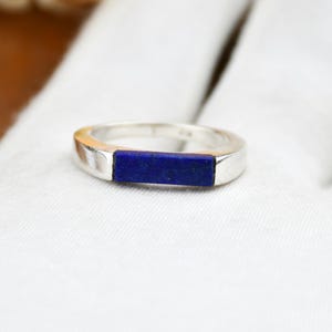 Op de afbeelding: Een zilveren ring met een rechthoekige, diepblauwe lapis lazuli steen in het midden. De ring wordt getoond op een witte ondergrond, wat het contrast tussen het metaal en de steen benadrukt.