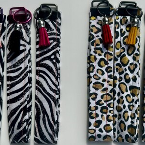 May include: A collection of wrist keychains with various patterns and colors. The keychains feature black and white zebra stripes and leopard print designs. Each keychain has a metal ring and a tassel in different colors.