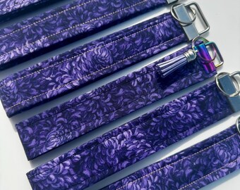 May include: Several purple fabric key fobs with a floral pattern. Each fob has a silver-colored metal ring and a matching fabric loop. One fob has a small, purple tassel and a rainbow-colored metal ring.