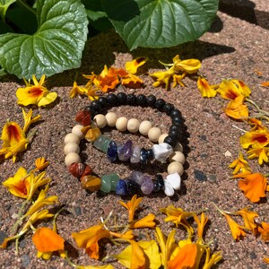 Op de afbeelding: Twee armbanden met kralen en natuurstenen accenten. Eén armband heeft zwarte lavakralen, terwijl de andere houten kralen combineert met kleurrijke edelstenen. De armbanden worden gepresenteerd op een getextureerd oppervlak, omringd door gele en oranje bloemen en groene bladeren.