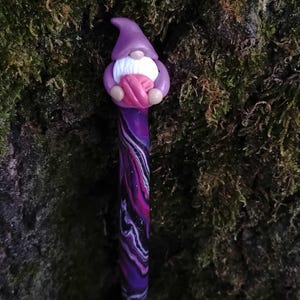May include: A purple and black crochet hook with a gnome holding a pink ball of yarn at the top. The gnome has a purple hat and white beard.