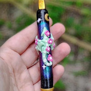 Puede incluir: Un ganchillo morado y negro con un dise&ntilde;o de galaxia y un adorno floral verde y rosa. El ganchillo es de 6,0 mm.