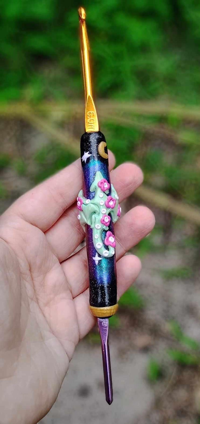 Puede incluir: Un ganchillo morado y negro con un dise&ntilde;o de galaxia y un adorno floral verde y rosa. El ganchillo es de 6,0 mm.
