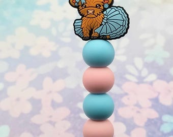 May include: A stack of blue and pink silicone beads with a Highland cow charm wearing a blue tutu and a bow on top.