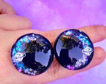 Puede incluir: Un par de pendientes de resina negra con temática de galaxia. Los pendientes presentan un fondo de purpurina plateada con estrellas blancas y un planeta plateado con anillos.