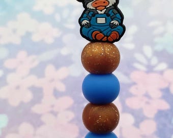 May include: A blue and orange cow charm with a white hat and a brown and gold bead necklace. The charm is sitting on top of a blue bead.