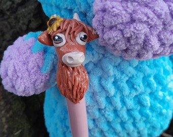 May include: A blue and purple crocheted plush toy with a brown and white cow head attached to it. The cow head has a sunflower on top and is made of polymer clay.