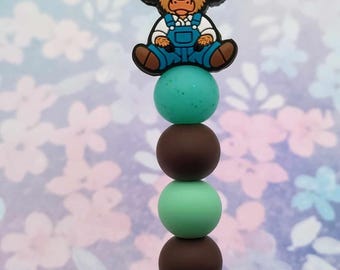 May include: A colorful beaded keychain with a cartoon highland cow wearing a red baseball cap and blue overalls. The beads are brown and teal green.