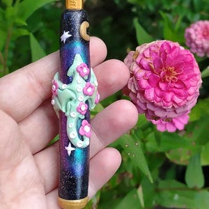 Puede incluir: Un gancho de ganchillo de 6,0 mm con un mango de tem&aacute;tica gal&aacute;ctica que presenta un drag&oacute;n verde con flores rosas y estrellas blancas. El mango es negro con un brillo iridiscente morado y azul.