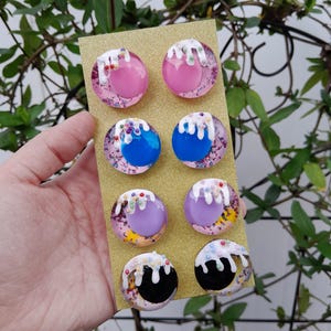 May include: Eight colorful, round earrings on a gold card. The earrings feature pink, blue, purple, and black centers with white icing and sprinkles. The background has a glittery effect.