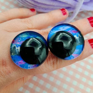 May include: Two large, round, glass eye cabochons with a black pupil and a blue, pink, and white galaxy design. The cabochons are held in a hand with red nail polish.