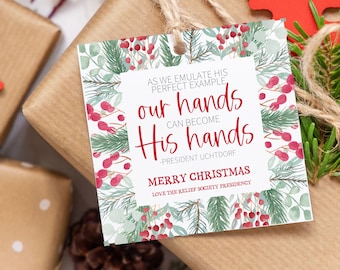May include: A white Christmas gift tag with a watercolour design of green pine branches and red berries. The text reads "As we emulate His perfect example, our hands can become His hands -President Uchtdorf Merry Christmas Love the Relief Society Presidency".