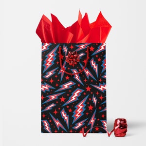 Pode incluir: Saco de presente preto com um padrão de raios e estrelas vermelhas, brancas e azuis. O saco tem uma alça vermelha e é preenchido com papel de seda vermelho. Uma fita e um laço vermelhos também estão presentes.