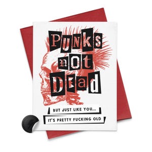 Op de afbeelding: Een witte wenskaart met een rode envelop toont een rood-zwarte schedelillustratie met de tekst "Punks Not Dead". Onder de schedel staat op de kaart "BUT JUST LIKE YOU... IT'S PRETTY FUCKING OLD". Links zit een zwarte sticker met een bliksemschicht.