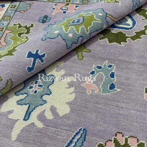 May include: A rolled-up, intricately patterned rug in shades of lavender, blue, green, and pink. The rug features a repeating floral and geometric design. The text "Rizwan Rugs" is visible on the rug's surface.