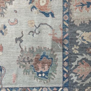 May include: A close-up of a rug with a light beige background and a blue, green, and red floral pattern. The rug has a dark blue border with a geometric pattern.