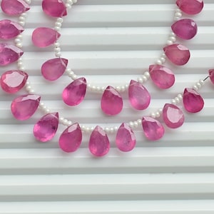 Può includere: Tre fili di perline di pietre preziose rosa con taglio a goccia, infilate su un filo di perline bianche. Le pietre preziose sono di un colore rosa vibrante e sono esposte su una superficie bianca e scanalata. Una nappa rossa è attaccata a un'estremità.