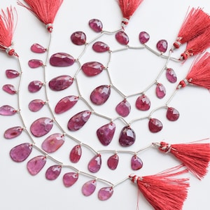 Pode incluir: Um fio de contas de pedras preciosas cor-de-rosa com conectores prateados e borlas vermelhas em cada extremidade. As contas são de forma oval e têm um acabamento facetado.
