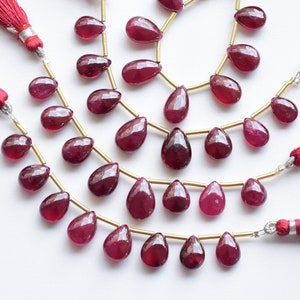Puede incluir: Una hebra de cuentas de piedras preciosas de color rojo rubí enhebradas en un alambre dorado con borlas rojas en cada extremo. Las cuentas tienen forma de pera y tienen un acabado brillante.
