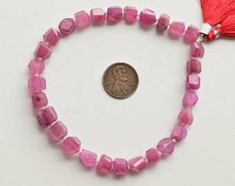 May include: A strand of pink gemstone beads, with a faceted, square shape. The beads are strung on a white cord and are approximately the size of a US penny.