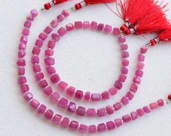 May include: Strands of faceted, cube-shaped, pink gemstones. The beads are strung together with a silver-colored string. Red tassels are attached to the ends of the strands. The gemstones have a polished, reflective surface.