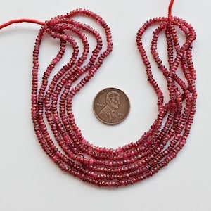 Puede incluir: Una hebra de cuentas de rubí rojo, de aproximadamente 2 mm de tamaño, con un borla en un extremo. Las cuentas son facetadas y tienen un acabado brillante. La hebra se coloca en un círculo con una moneda de un céntimo de euro para la escala.