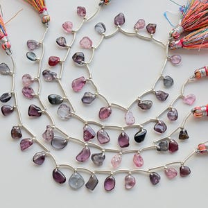 May include: A strand of faceted gemstone beads in shades of pink, purple, and blue, strung on silver wire with small silver spacers. The beads are teardrop shaped and have a natural, irregular look. The strand is finished with colorful tassels.