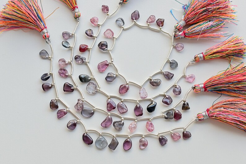 May include: A strand of faceted gemstone beads in shades of pink, purple, and blue, strung on silver wire with small silver spacers. The beads are teardrop shaped and have a natural, irregular look. The strand is finished with colorful tassels.