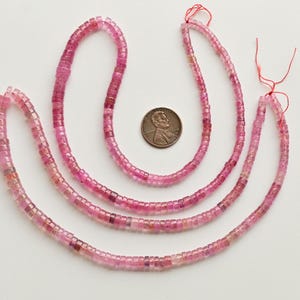 May include: Three strands of pink tourmaline beads, each strand is approximately 18 inches long. The beads are faceted and have a square shape. The beads are strung on a clear thread and are separated by small knots. The beads are arranged in a U-shape and are displayed on a white surface.