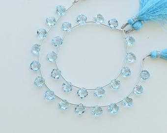 May include: Two strands of light blue gemstone beads with faceted, flower-shaped stones. The strands are strung on silver wire and finished with matching blue tassel accents. The beads are displayed on a white surface.