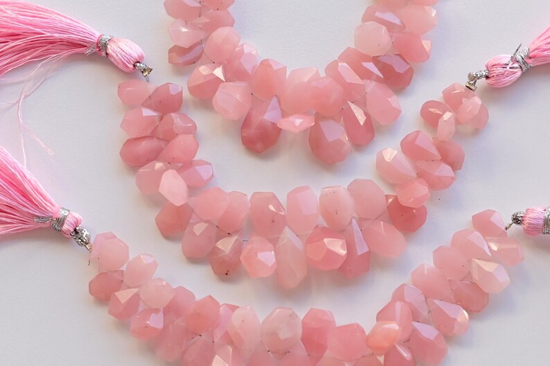 May include: Three strands of pink rose quartz gemstone beads, each strand is approximately 18 inches long and features faceted, teardrop-shaped beads. The beads are strung on a silver wire and have a tassel at each end.