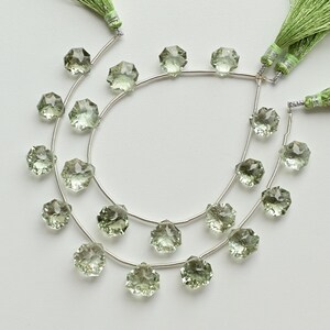 May include: Two strands of faceted, pale green gemstone beads on silver wire, with green tassel accents. The beads have a multi-faceted, star-like shape. The background is a plain white surface. Ideal for jewelry making.