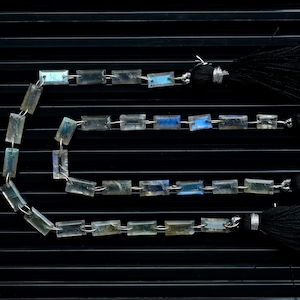 May include: Three strands of rectangular, iridescent gemstone beads are displayed against a black background. Each strand is connected to a black tassel with silver-colored metal accents. The gemstones have a mix of blue, green, and clear tones.