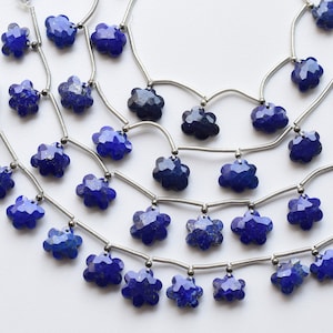 May include: A strand of blue lapis lazuli gemstone beads in a flower shape, strung on silver wire with silver spacers. The beads are faceted and have a sparkly finish. The strand is finished with white tassels on each end.