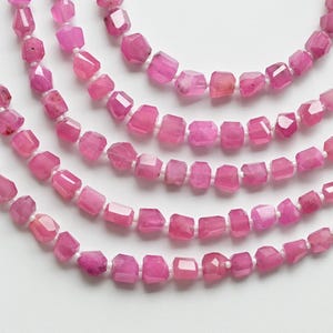 May include: A strand of pink gemstone beads, faceted and strung on white thread. The beads are square-shaped and have a slightly irregular, natural look.