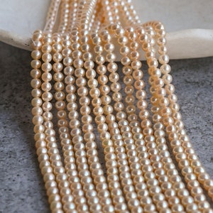 May include: A strand of cream-colored pearl beads, arranged in rows, on a white surface. The pearls are round and smooth, and they have a subtle sheen.