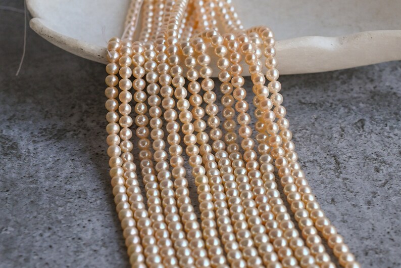 May include: A strand of cream-colored pearl beads, arranged in rows, on a white surface. The pearls are round and smooth, and they have a subtle sheen.