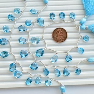 May include: A strand of light blue topaz gemstone beads strung on silver wire with silver bead accents. The beads are faceted and have a teardrop shape. The strand is finished with blue tassel charms.